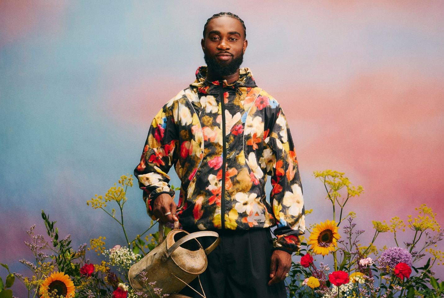 Kofi Stone wearing a floral jacket surrounded by flowers holding a watering can